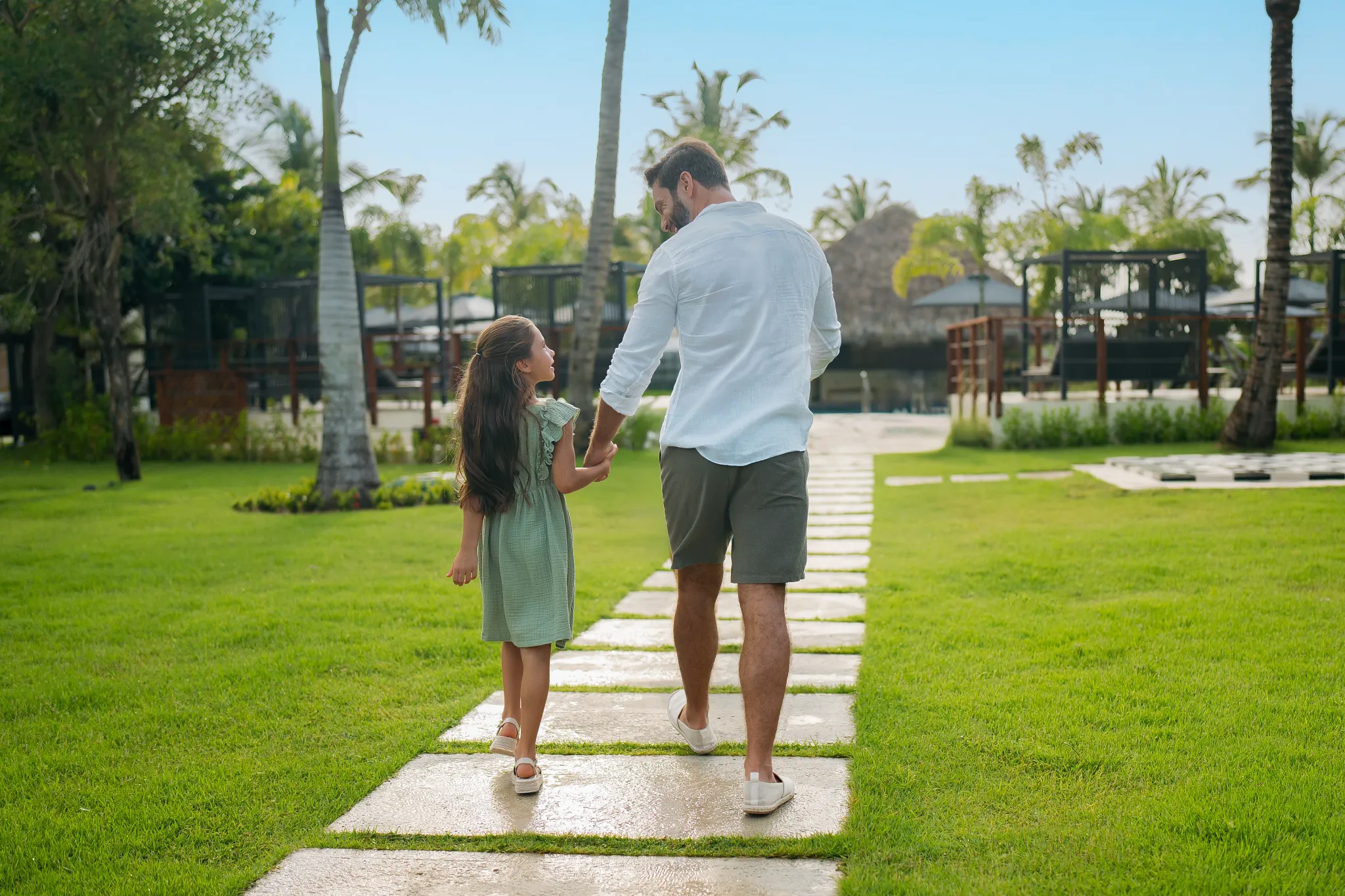 Father and daughter walking together along a garden pathway at Dreams Royal Beach Punta Cana surrounded by palm trees and greenery.