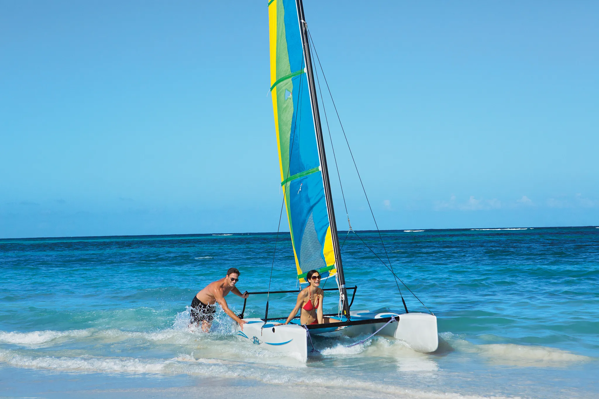 Couple pushing a Hobie Cat sailboat into the ocean at Dreams Royal Beach Punta Cana, enjoying beachfront water sports.